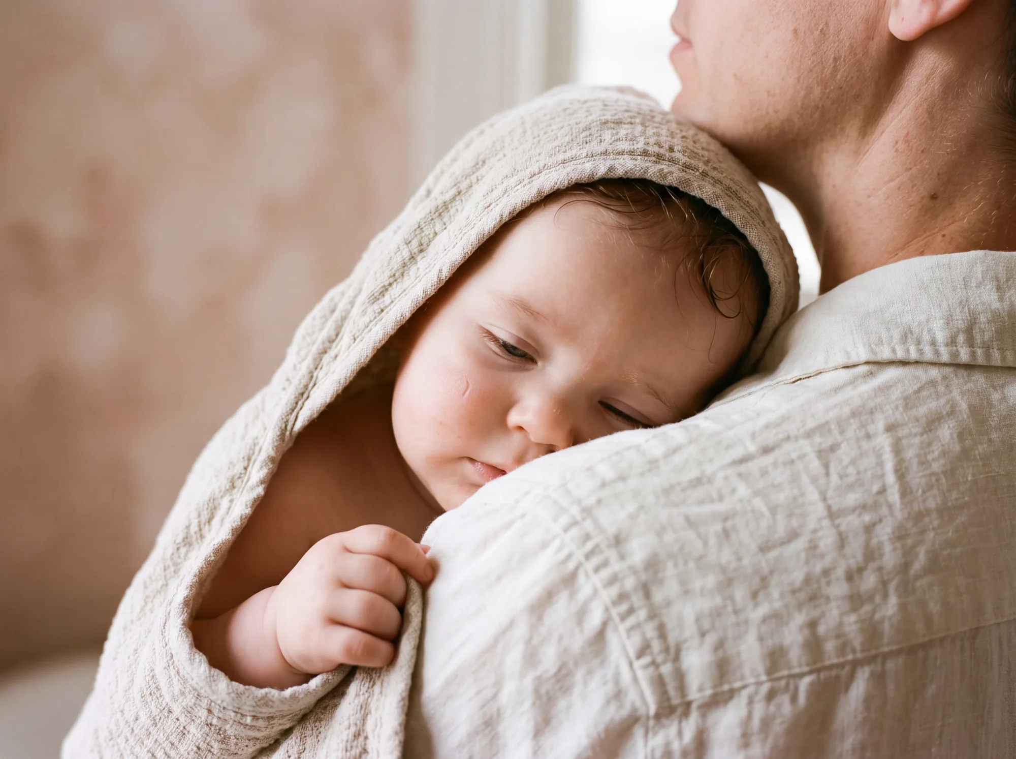 A sleepy baby wrapped in a hooded towel on a parent’s shoulder after the bath.